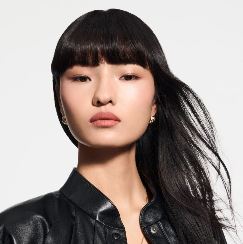 Close-up portrait of an Asian woman with long black hair and bangs, wearing a black leather jacket against a light background.