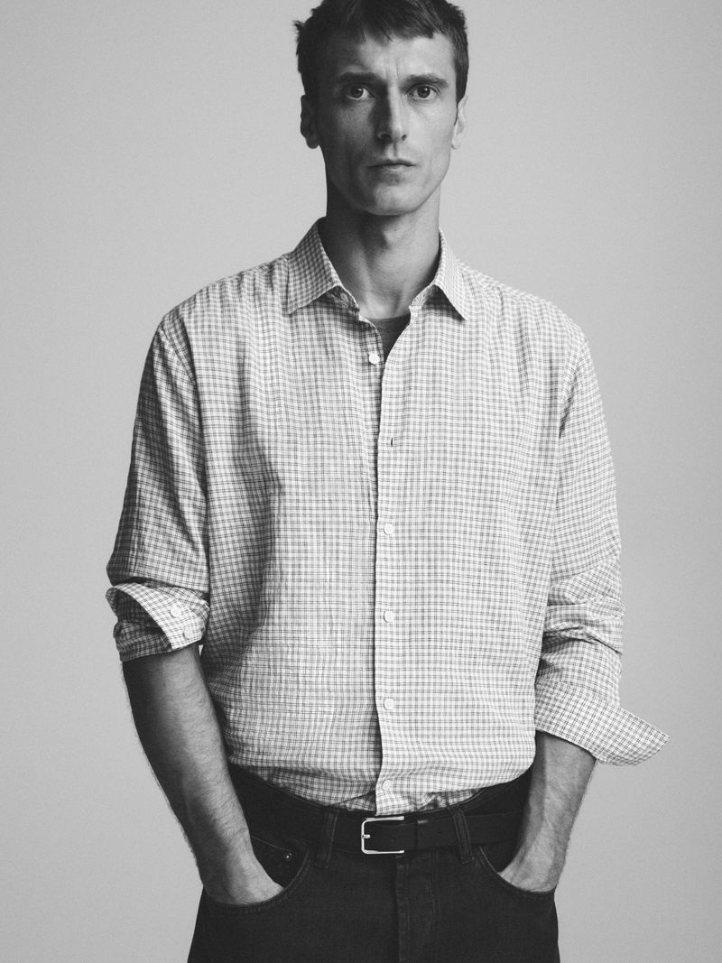 Black and white portrait of an adult man in a button-down shirt and jeans, standing indoors with a neutral expression and studio lighting.
