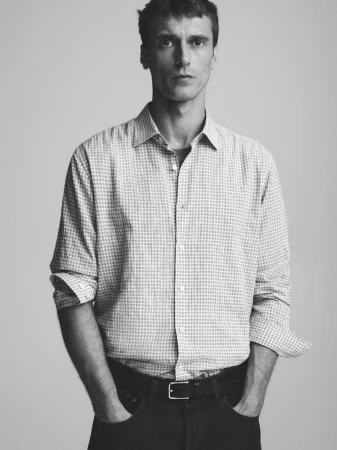 Black and white portrait of an adult man in a button-down shirt and jeans, standing indoors with a neutral expression and studio lighting.