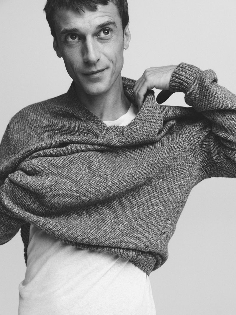 Monochrome portrait of a man in a gray knit sweater, smiling slightly, indoors against a neutral background. Fashion and portraiture.