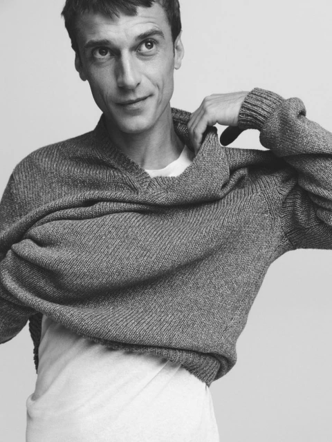 Monochrome portrait of a man in a gray knit sweater, smiling slightly, indoors against a neutral background. Fashion and portraiture.