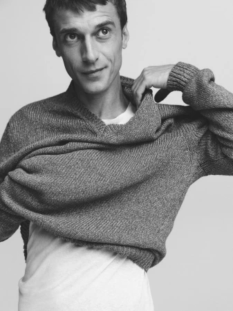 Monochrome portrait of a man in a gray knit sweater, smiling slightly, indoors against a neutral background. Fashion and portraiture.