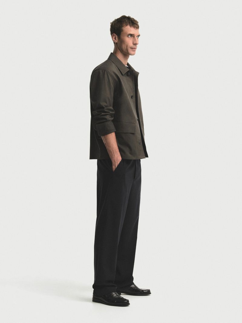 A man in a green jacket and dark pants stands in a brightly lit studio with a clean, neutral background, looking to the side.
