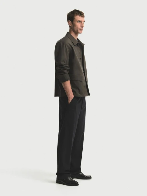 A man in a green jacket and dark pants stands in a brightly lit studio with a clean, neutral background, looking to the side.