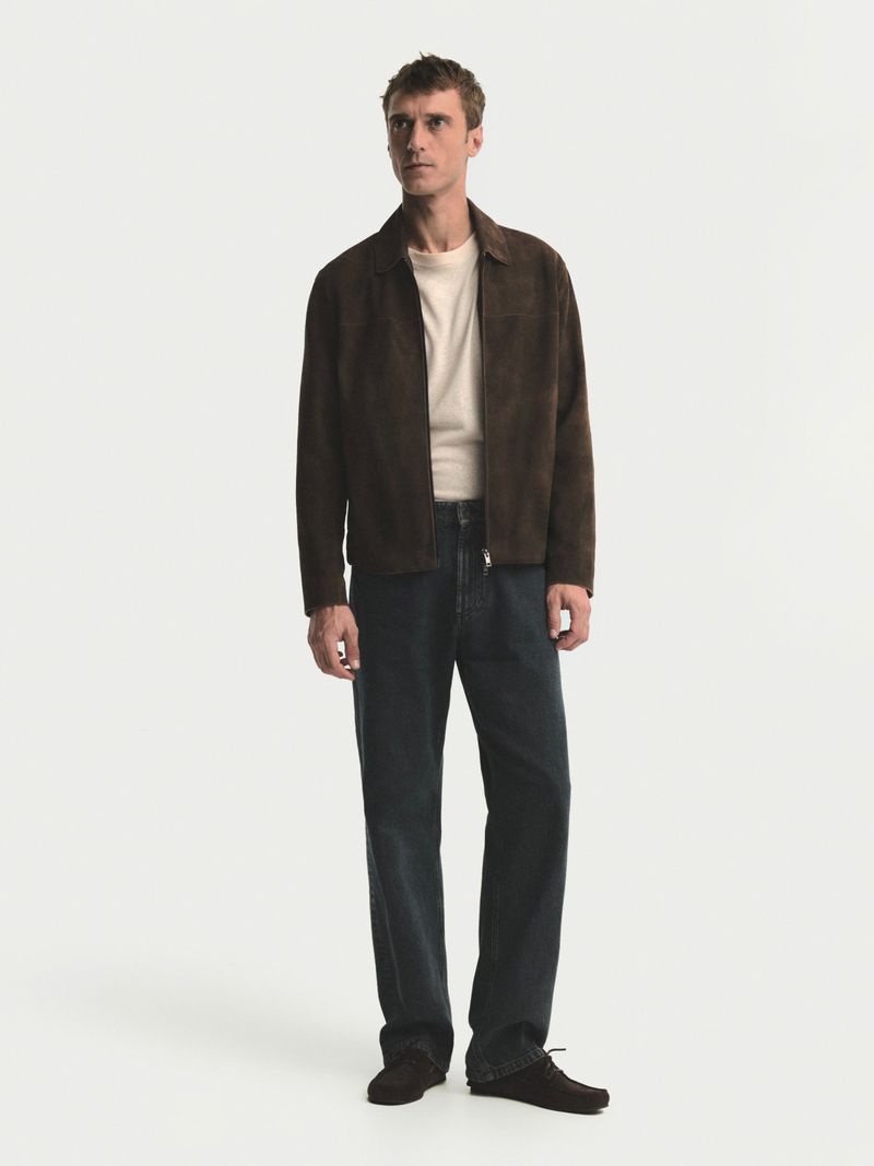 A full-length shot of a male model showcasing a brown suede jacket paired with a cream-colored tee and dark denim pants against a clean white backdrop.