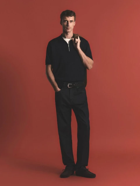 Full-length studio shot of an adult male in a black polo shirt and black pants, standing in a neutral pose against a red background.