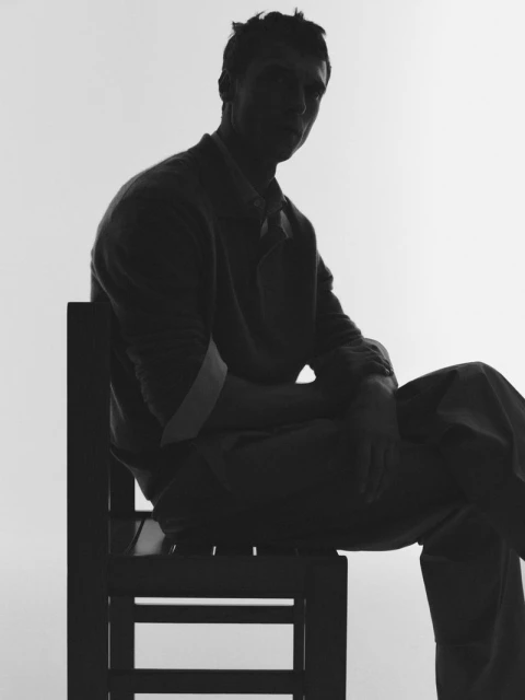 Silhouette of a man sitting on a chair, wearing a sweater and collared shirt against a white background in a studio-style setting.