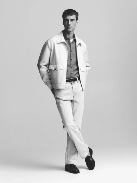 Monochrome portrait of a man in light jeans and jacket, leaning with crossed legs, set against a neutral background in a fashion style.