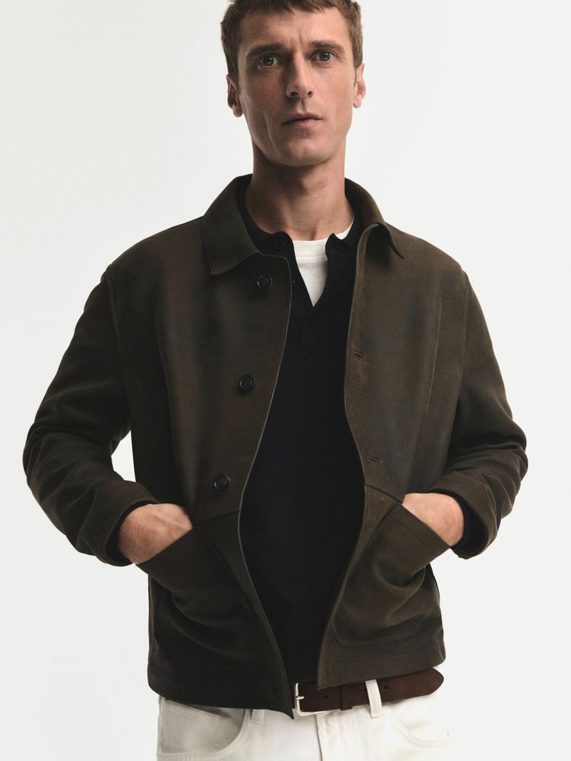 Man in olive green jacket with hands in pockets, black shirt, and white trousers against a clean, neutral background in a studio setting.