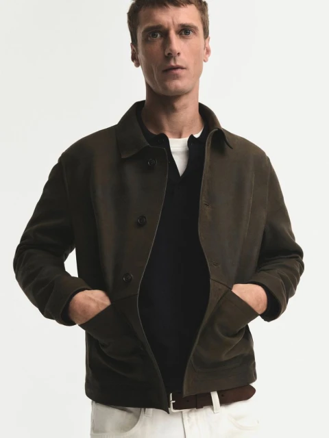 Man in olive green jacket with hands in pockets, black shirt, and white trousers against a clean, neutral background in a studio setting.