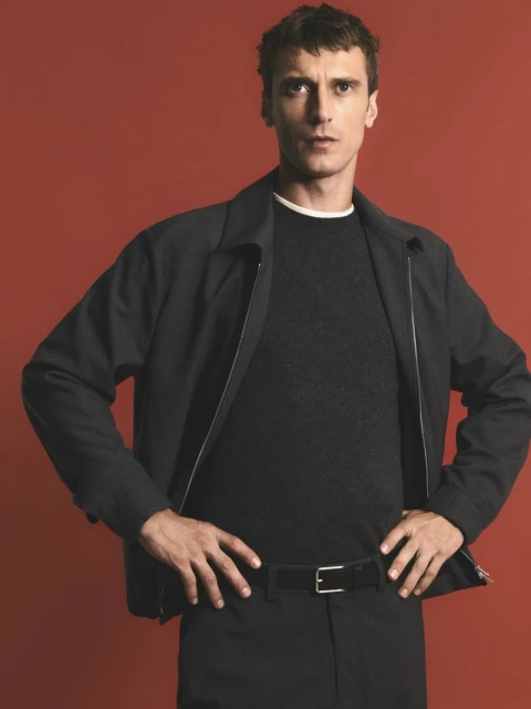 Studio shot of a man in black jacket and pants, hands on hips, against a muted red background. Fashion editorial with clean aesthetic.