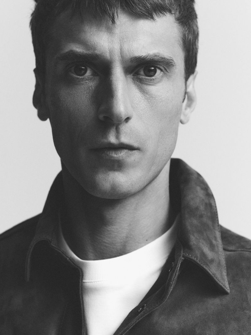 Portrait of a man with short dark hair in a close-up studio shot, wearing a suede jacket over a white crewneck shirt on white background.