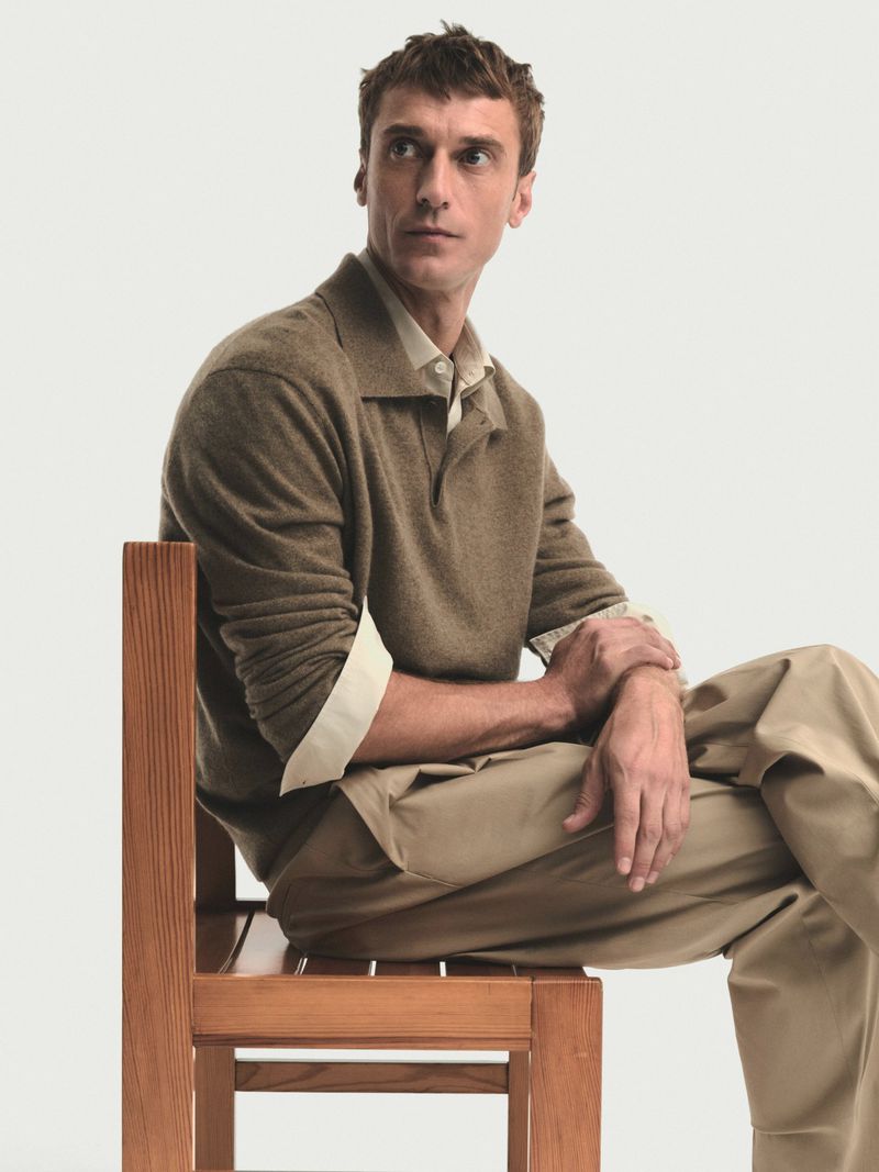 Man sits on a wooden chair in front of a white backdrop, wearing khaki pants, a light-colored shirt, and a brown collared sweater.