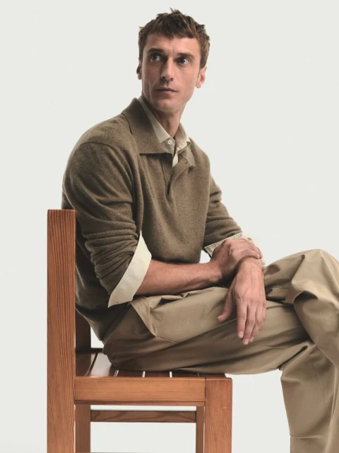 Man sits on a wooden chair in front of a white backdrop, wearing khaki pants, a light-colored shirt, and a brown collared sweater.