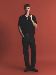 Full-length portrait of a male model wearing a black zip polo shirt and trousers against a warm terracotta backdrop.