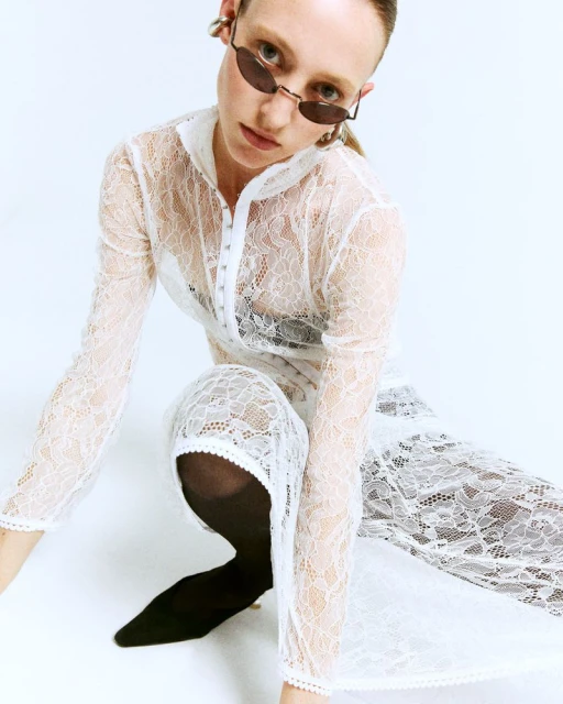 Woman in a white lace dress and sunglasses poses in a photo studio. The indoor shot captures fashion and studio lighting.