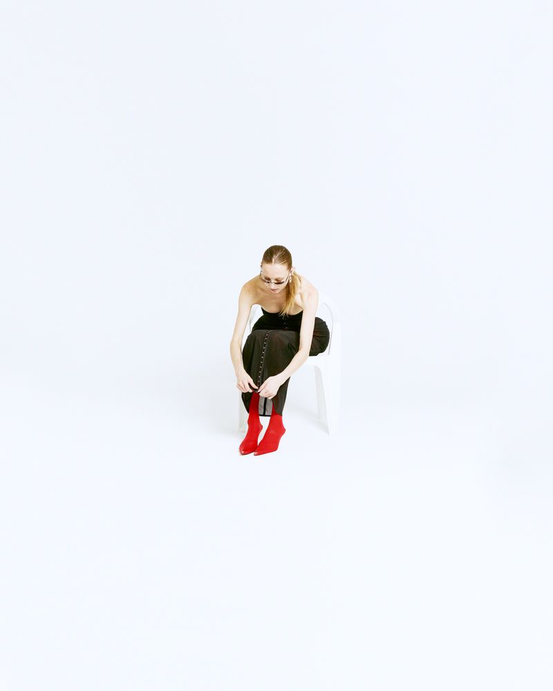 Woman sits on a chair in a photo studio, tying the laces of her red boots. She wears sunglasses and black top with leggings.