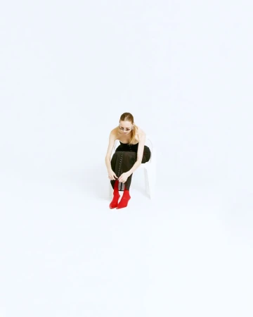 Woman sits on a chair in a photo studio, tying the laces of her red boots. She wears sunglasses and black top with leggings.