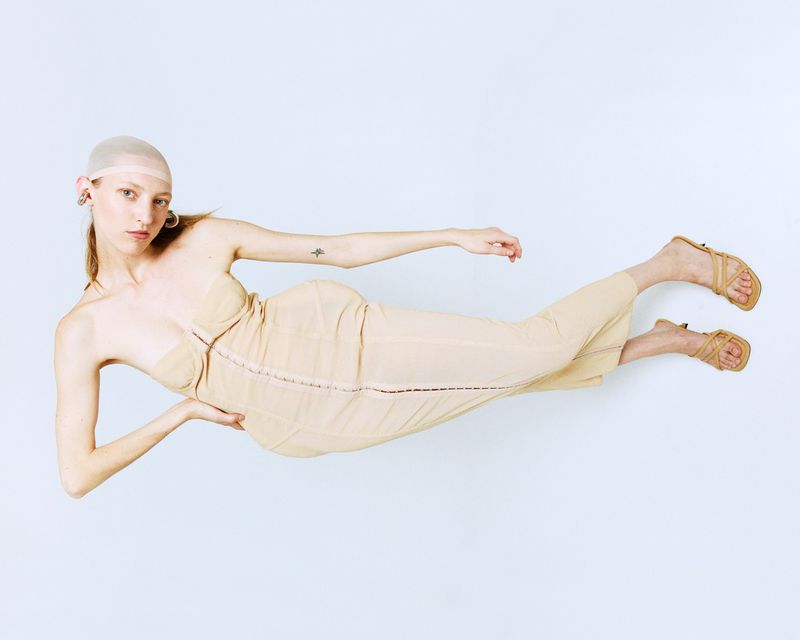 Woman in tan dress and head covering poses on white background. Fashion shot with long legs extended, neutral facial expression.