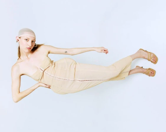 Woman in tan dress and head covering poses on white background. Fashion shot with long legs extended, neutral facial expression.