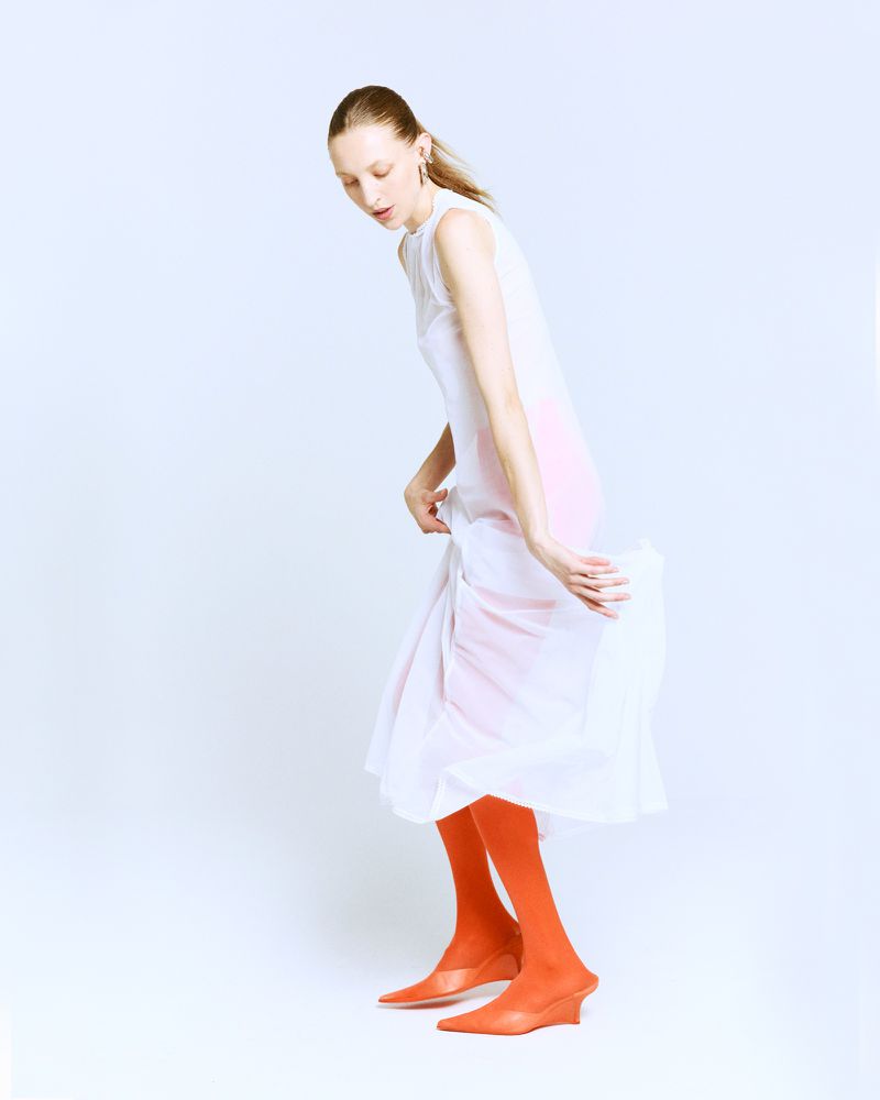 A slender woman poses in a flowing white dress with orange tights and heels against a bright white backdrop in a fashion shot.