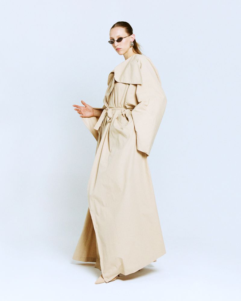 A slender female model strikes a pose in a full-length beige trench coat and sunglasses against a bright white background, studio setting.