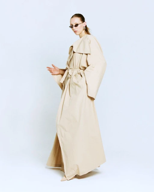 A slender female model strikes a pose in a full-length beige trench coat and sunglasses against a bright white background, studio setting.
