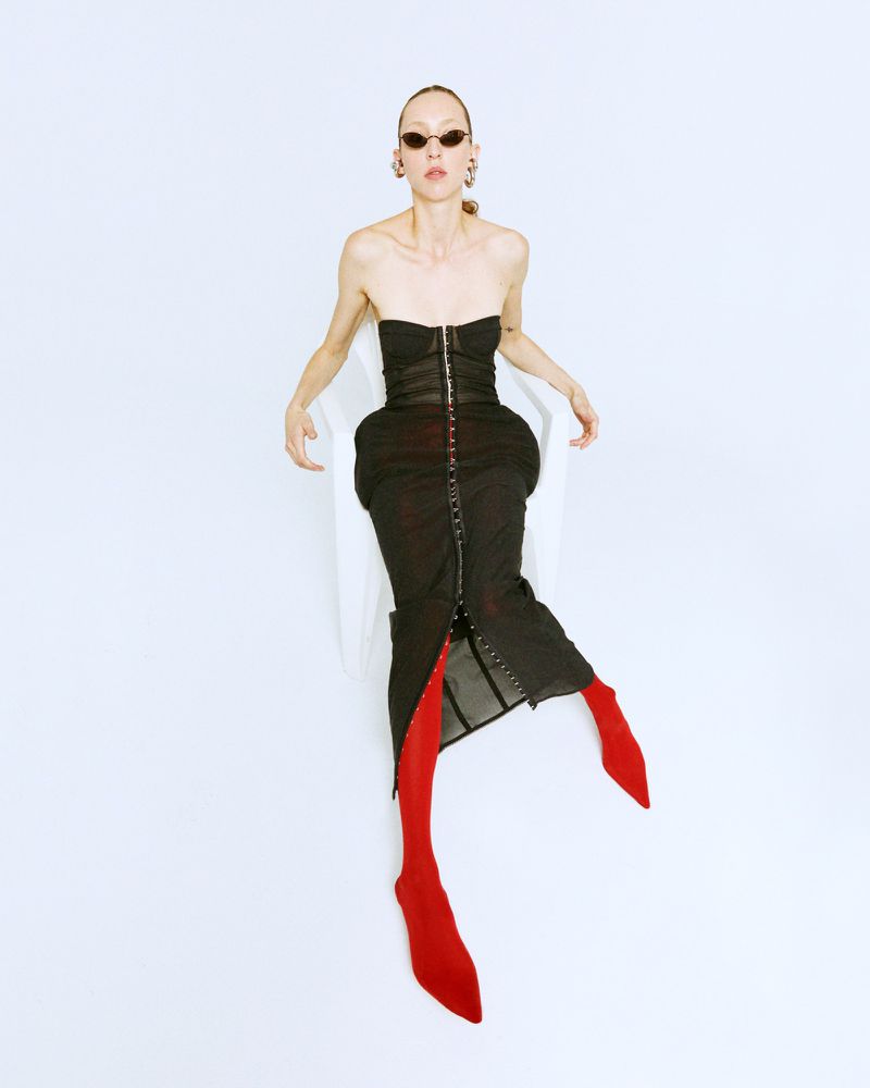 A slender woman in a black bustier dress and red tights sits in a white chair against a white backdrop, showcasing a fashion-forward studio shot.