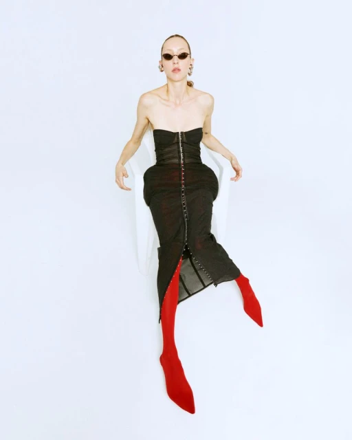 A slender woman in a black bustier dress and red tights sits in a white chair against a white backdrop, showcasing a fashion-forward studio shot.