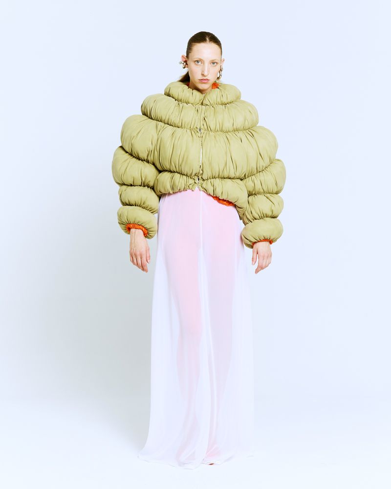Model wearing an oversized sculptural green puffer jacket and sheer floor-length skirt against a white studio background.