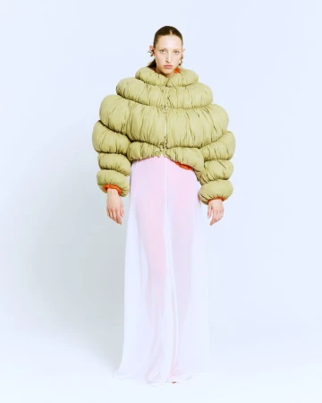Model wearing an oversized sculptural green puffer jacket and sheer floor-length skirt against a white studio background.