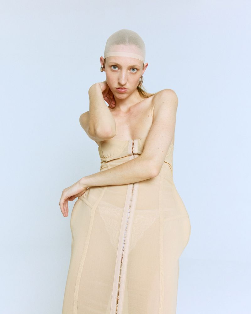 Portrait of a woman wearing a beige corset dress and skin-colored cap, posing against a light blue backdrop in a studio setting.
