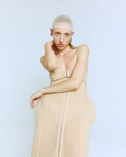 Portrait of a woman wearing a beige corset dress and skin-colored cap, posing against a light blue backdrop in a studio setting.