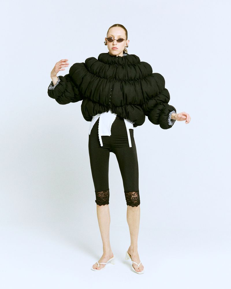 Fashion photo of a slender adult woman wearing a black puffer jacket, black lace leggings, white sandals, and oval sunglasses in a white studio.