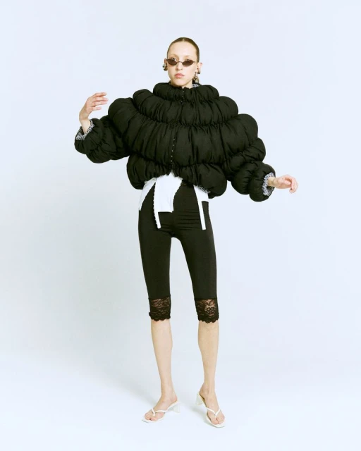 Fashion photo of a slender adult woman wearing a black puffer jacket, black lace leggings, white sandals, and oval sunglasses in a white studio.