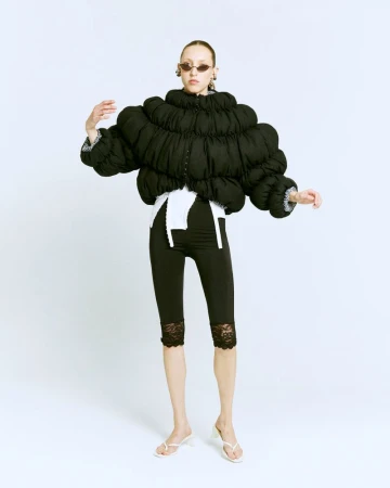 Fashion photo of a slender adult woman wearing a black puffer jacket, black lace leggings, white sandals, and oval sunglasses in a white studio.