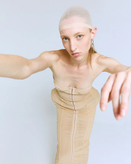A fair-skinned model wearing a nude corset-style dress and a nylon cap poses in a bright, direct, full studio shot on a white background.