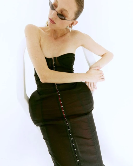 An adult female wearing a strapless black dress and sunglasses leans against a white chair in a studio setting with soft lighting.