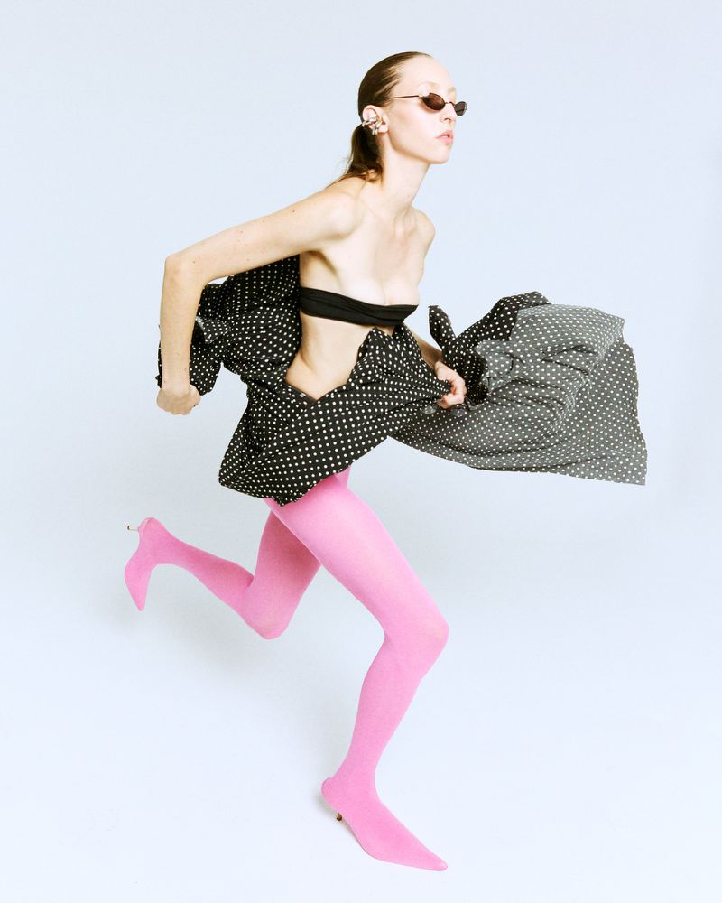 Fashion model in motion wearing a black top, polka dot skirt, pink tights, and sunglasses, posing against a white backdrop in a studio.