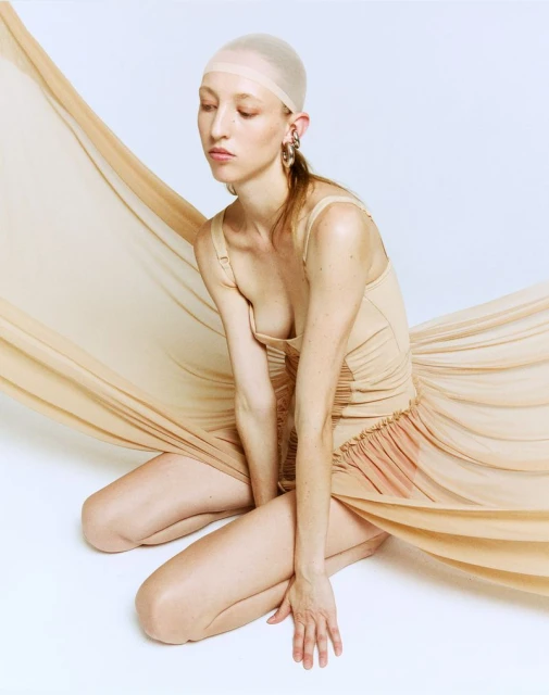 Woman in nude bodysuit and stocking cap kneeling indoors with tan fabric draped around her against a white backdrop in a fashion shot.