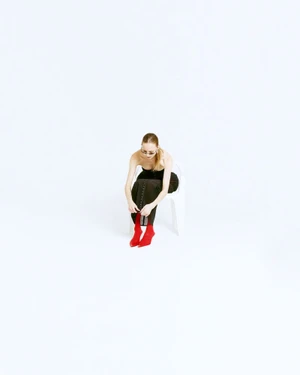 A woman wearing a dark strapless dress and red boots sits on a white chair against a bright white background.