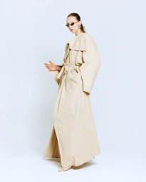 Model wearing a long beige belted trench coat and sunglasses against a plain white studio background.