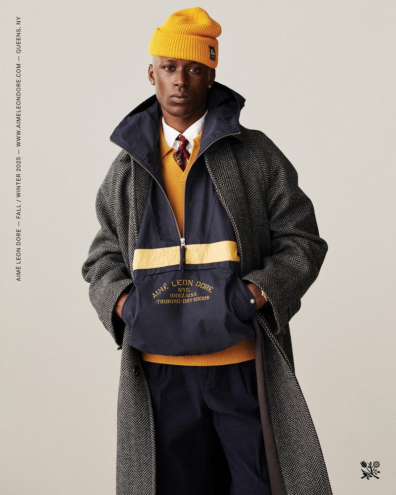 Man in long herringbone coat, yellow beanie, and navy anorak stands against a neutral backdrop in a fashion editorial image.