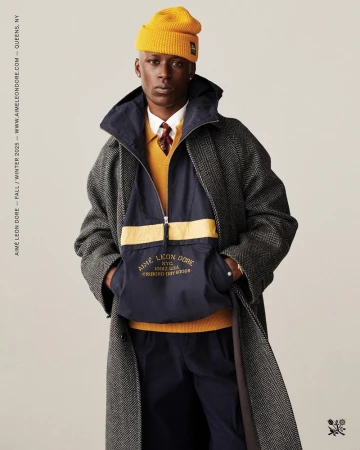 Man in long herringbone coat, yellow beanie, and navy anorak stands against a neutral backdrop in a fashion editorial image.