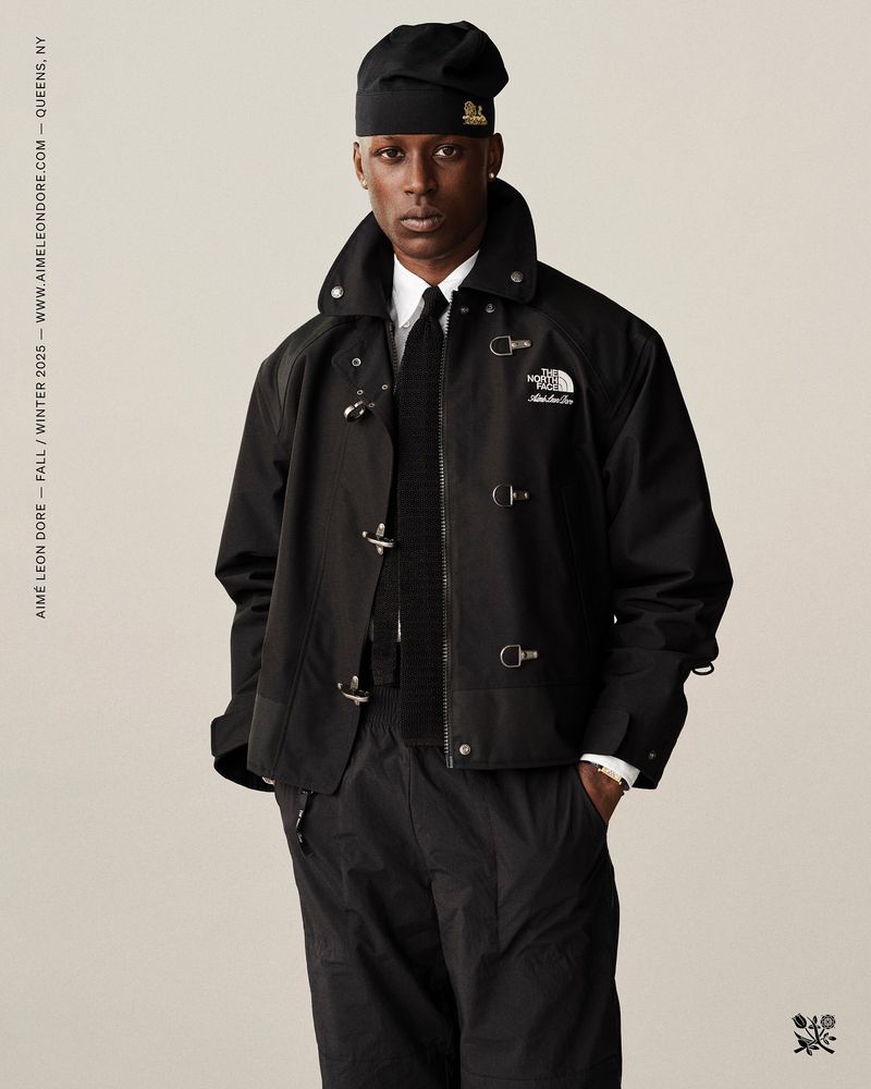 A Black male fashion model poses in an outdoor studio wearing a black jacket, matching pants, and a beanie against a neutral background.