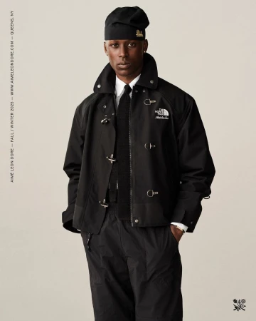 A Black male fashion model poses in an outdoor studio wearing a black jacket, matching pants, and a beanie against a neutral background.