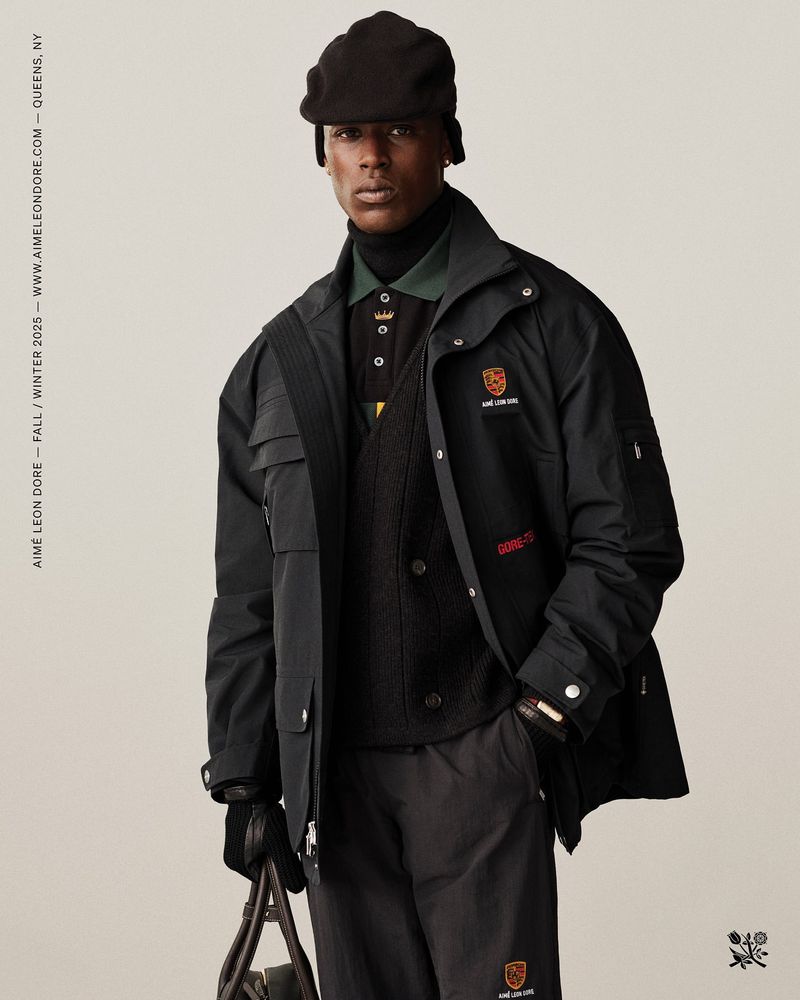 A black male fashion model poses in a black jacket, trousers, and cap against a cream background, holding a bag in his gloved hand.