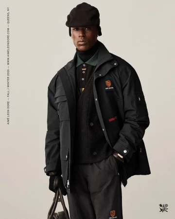 A black male fashion model poses in a black jacket, trousers, and cap against a cream background, holding a bag in his gloved hand.