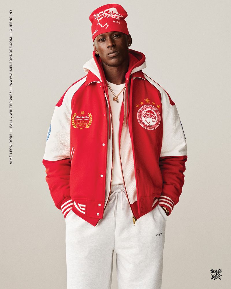 An African-American man is posing in a red and white jacket, grey sweatpants, and a red knit hat, standing in a neutral studio.