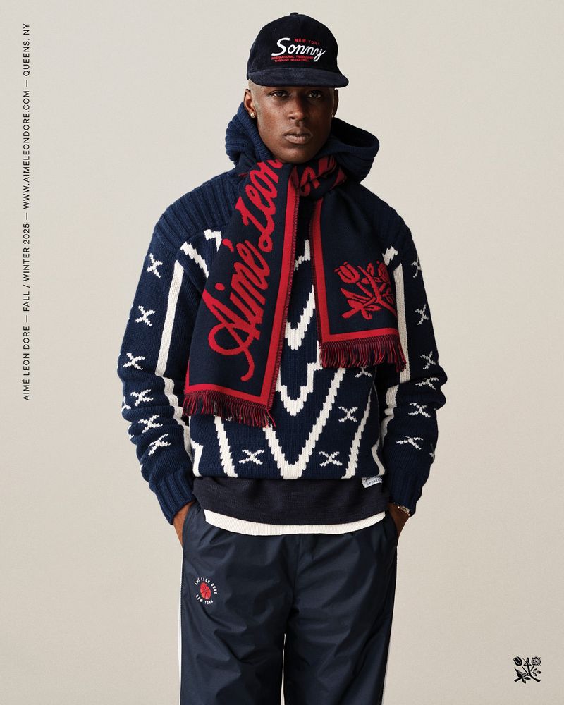 Man in a blue and white patterned sweater, blue pants, scarf, and hat stands against a neutral backdrop in a studio shot.